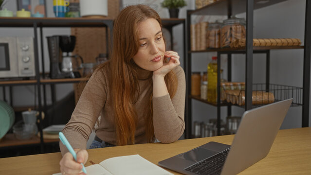 Woman working on laptop in cozy indoor office setting, featuring attractive redhead female surrounded by shelves, kitchenware, and sleek modern workspace ambiance.