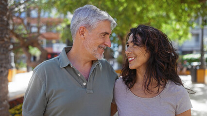 Middle-aged couple smiling lovingly at each other in an outdoor urban park setting, highlighting their beautiful relationship and shared happiness in the warm daytime light.