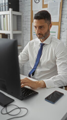 Obraz premium Businessman working intently at computer in modern office, wearing formal shirt and tie, surrounded by files and documents, suggesting professional environment and productivity.
