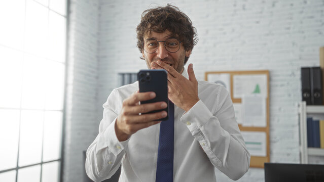 Young man in an office smiles at his smartphone displaying a joyful expression in a bright modern workspace with white walls and pinned documents.
