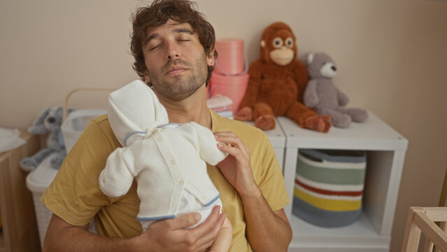 Young man holding baby in arms sitting in cozy bedroom surrounded by toys with a serene expression showcasing fatherhood and domestic life indoors.