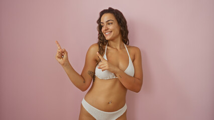 Naklejka premium Young, attractive, hispanic woman in a white bikini pointing sideways against a pink background, showcasing beauty and confidence with a smile