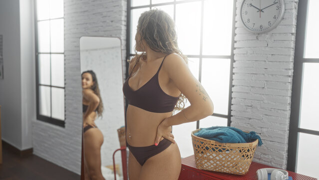Young, attractive, brunette woman in lingerie admiring her reflection in a bedroom mirror with a basket of blue towels and a wall clock in the background