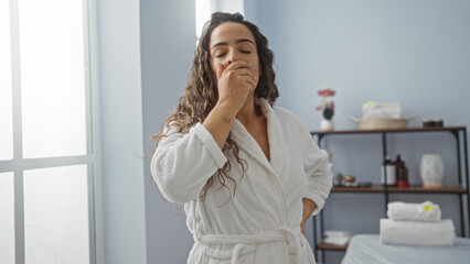 Fototapeta premium Woman yawning in a spa room while wearing a white robe, surrounded by a serene interior, showcasing relaxation in a wellness center.