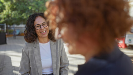 Women laughing outdoors in an urban park setting, showing friendship and happiness in a city environment, with two middle-aged friends engaging in joyful conversation.