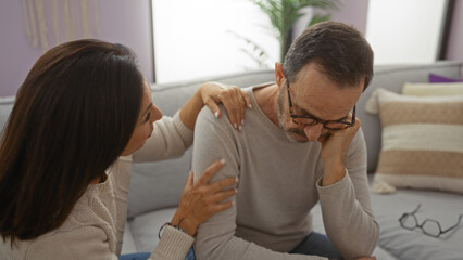 Fototapeta premium Middle aged hispanic couple sitting together in a living room with woman comforting man, highlighting love and family in an intimate indoor setting.