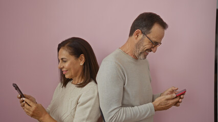 Middle-aged hispanic couple stands back-to-back using smartphones, woman in white, man in gray, over pink background wall expressing love and connection.