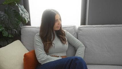 Obraz premium Woman sitting on cozy couch in living room with plants and soft pillows, reflecting a peaceful and relaxed home atmosphere with warm ambient light.