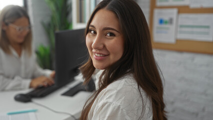 Obraz premium Woman smiling in an office environment with a coworker working in the background, highlighting a collaborative workplace atmosphere indoors.
