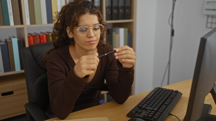 Young woman wearing glasses sitting at a desk in an office room holding a pen while looking...