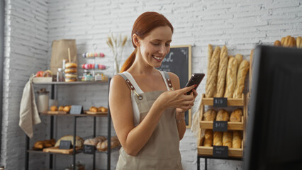 Young woman smiling and using phone in bakery shop with shelves of bread and pastries in the background