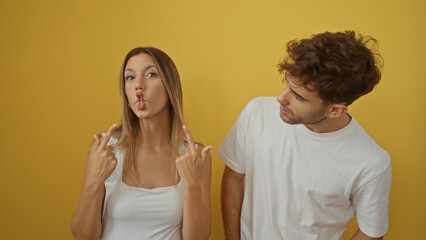 Woman making funny face gestures next to man smiling warmly both wearing white shirts in front of yellow background