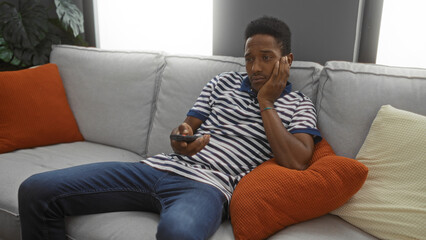 Obraz premium Young man lounging on sofa in cozy living room, holding remote, and appearing contemplative, surrounded by cushions and soft lighting.