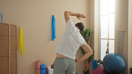 Obraz premium Man stretching in wellness room with yoga mats and exercise balls, bright interior with natural light, showcasing relaxation in a tranquil setting, reinforcing fitness routine.