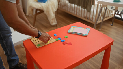 Man arranging colorful blocks on red table in cozy bedroom with baby's cradle in the background, highlighting a serene indoor family setting.
