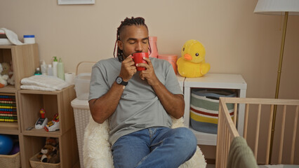 Young man with braids relaxing in a cozy bedroom, sipping from a red mug beside a baby's cradle and...