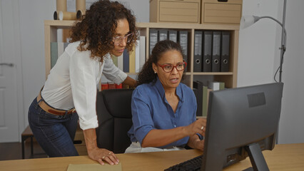 Hispanic women working together on a computer in an office environment, highlighting teamwork and collaboration in a professional setting.