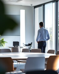 Fototapeta premium Businessman stands in modern office, looking out window at sunny cityscape.