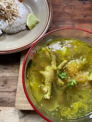 typical Indonesian food, chicken soto complete with claw and koya toppings, very suitable to be eaten with rice, boiled eggs, lime juice served in a glass bowl on a wooden table background