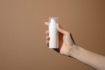 Woman's hand with manicured nails holds white bottle of cream. Unmarked container with transparent lid with pump. Background is plain, light brown in color, with slight hint of beige