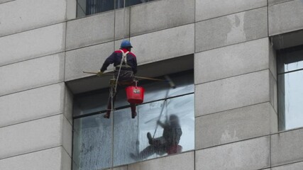 Workers clean high-rise windows in urban setting while suspended from ropes in bright conditions