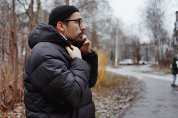 A young man in casual style walks through the snowy city on a winter day, chatting on his phone and...