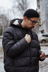 A young man in casual style walks through the snowy city on a winter day, chatting on his phone and...