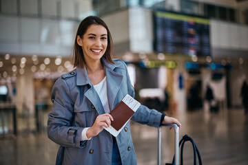 Happy traveler holding her passport at flight ticket at airport and looking at camera.