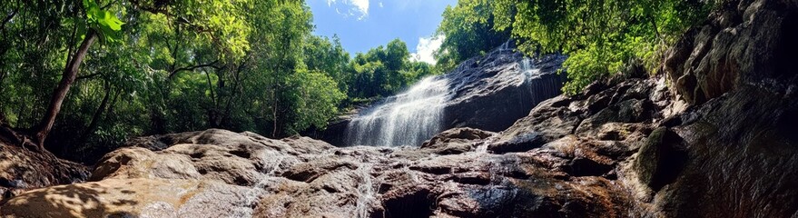 Fototapeta premium Majestic Waterfall Cascading Down Rocky Cliff Surrounded by Lush Green Forest and Bright Blue Sky