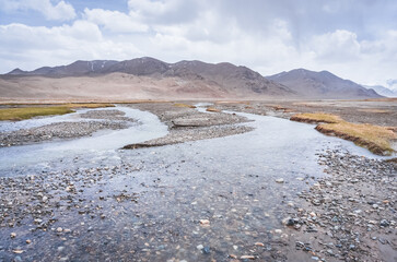 Mountain river flows in the valley in the highlands of Tien Shan in Pamir in Tajikistan, panoramic landscape for background in high mountains with rocks, snow and glaciers