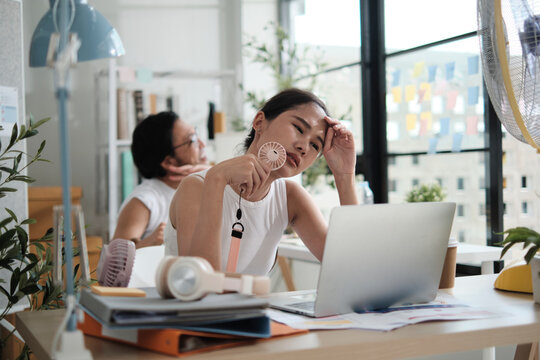 Two Asian workers are unhappy working with hot temperatures at small business office in summer season, uncomfortable with no air conditioner, using fans blowing wind, sitting at desks, and sweating.