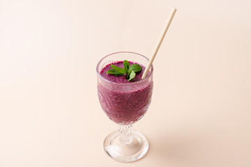 Cut glass cup with purple blueberry smoothie decorated with fresh mint leaves and a straw on a beige background. Copy Space. Healthy eating, antioxidants and wellness concept