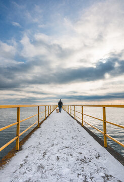 Man walking dog towards end of long pier on cold winter day.