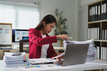 Young business woman in red suit working with a laptop and checking financial documents, managing...