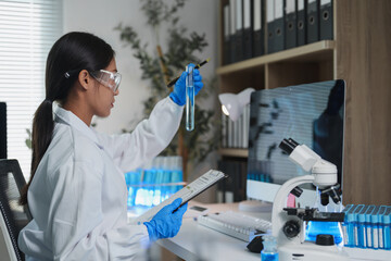 Female scientist wearing lab coat, safety glasses and gloves holding test tube with blue liquid and analyzing data on clipboard in modern laboratory with computer and microscope