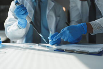 Two scientists in lab coats and blue gloves are conducting research, using a micropipette to...