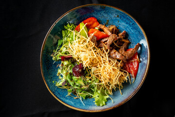 A vibrant plate featuring sliced beef stir-fried with bell peppers and onions, served alongside a fresh green salad topped with crispy fried potatoes, all presented on a decorative blue plate.