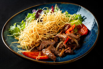 A vibrant plate featuring sliced beef stir-fried with bell peppers and onions, served alongside a fresh green salad topped with crispy fried potatoes, all presented on a decorative blue plate.