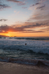 A vibrant sunset with orange and pink hues over a Hawaiian beach. Active ocean waves, a person in the water, and a distant land silhouette are visible.