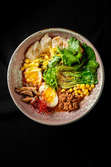 A beautifully arranged bowl of ramen featuring tender slices of meat, soft boiled eggs, fresh greens, corn, and vibrant garnishes, set against a dark background for contrast.
