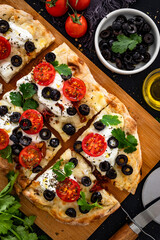 Pinsa Romana with burrata cheese, black olives and cherry tomatoes on black table	