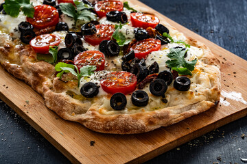 Pinsa Romana with burrata cheese, black olives and cherry tomatoes on black table	