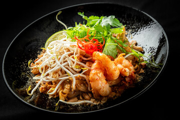 A vibrant plate of Pad Thai featuring shrimp, bean sprouts, fresh lime, peanuts, and cilantro, beautifully arranged on a dark background, highlighting its delicious textures and colors.