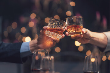 Whiskey Toast: Two hands in business attire raise glasses of amber whiskey in a celebratory toast against a softly blurred background of warm, festive lights.