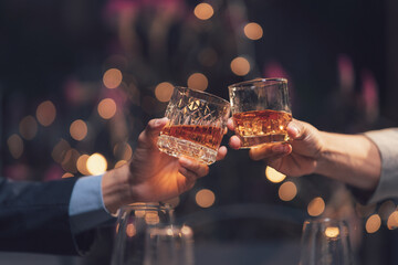 Toasting Success: Two hands in business attire raise glasses of amber liquid in a celebratory toast, set against a warm, bokeh-lit backdrop.  The image conveys partnership, success.