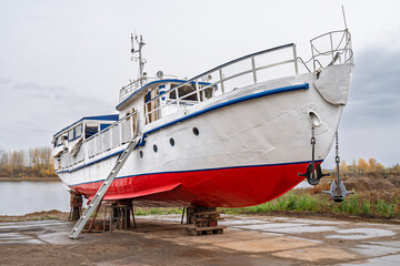 White and red ship raised on blocks for maintenance at dry dock near river, with cloudy sky and autumn trees in background. Concept of ship repair