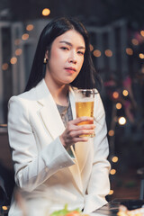 A Toast to the Weekend: A confident young woman in a crisp white blazer savors a glass of beer, her gaze fixed on the camera in a moment of quiet relaxation.