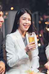 Cheers to Connection: A radiant young woman, dressed in a white blazer, raises a glass of beer with a joyful smile, enjoying a convivial gathering with friends.