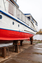 White and red ship raised on blocks for maintenance at dry dock near river, with cloudy sky and...