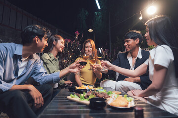 Nighttime Cheers: Friends gather around a table laden with food and drinks, raising their glasses in a toast, illuminated by warm lighting in a cozy outdoor setting.  